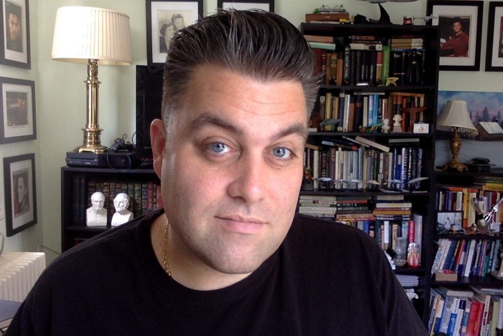 Smiling photo of poet and author, D. Ryan Lafferty in front of a overloaded bookshelf.